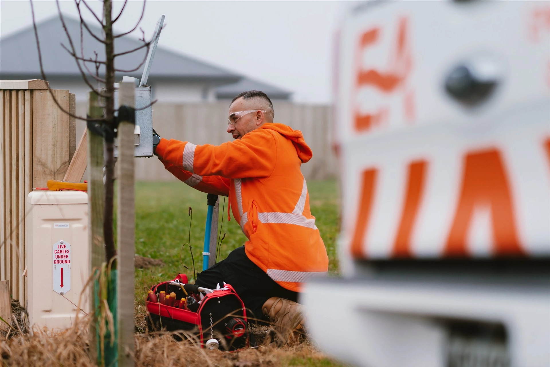 Worker in an orange jacket kneeling while inspecting an electrical box outdoors, with tools nearby and a blurred vehicle in the foreground.