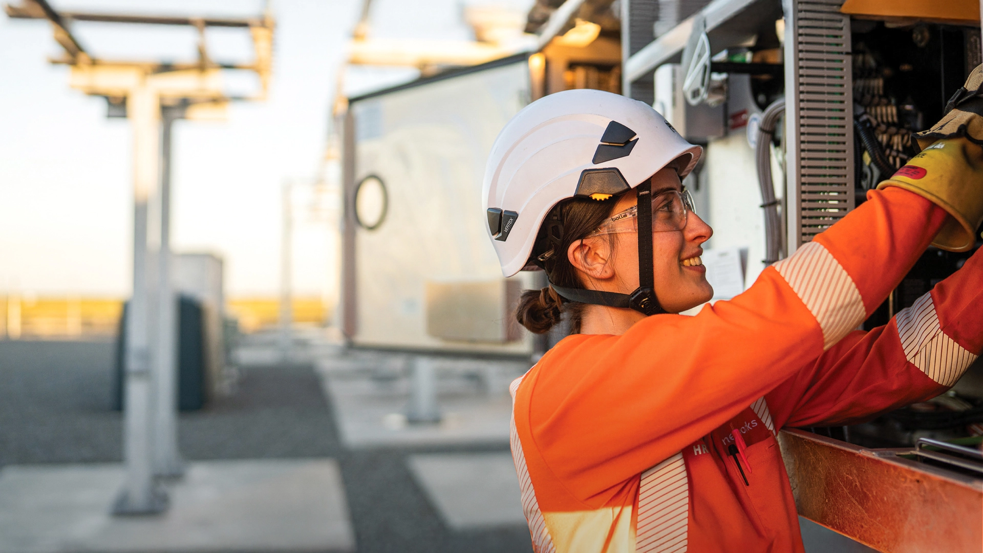 Worker in safety gear and helmet inspects electrical equipment outdoors, with solar panels in the background.